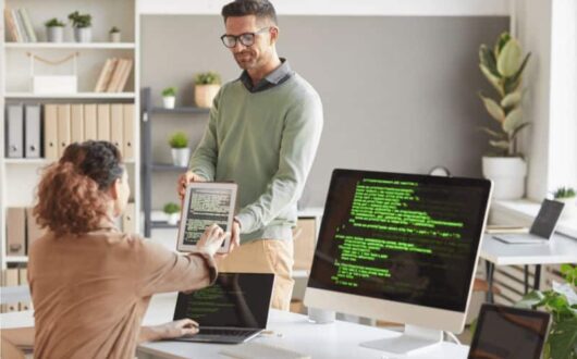 Une femme face à un écran de code échange avec un collaborateur